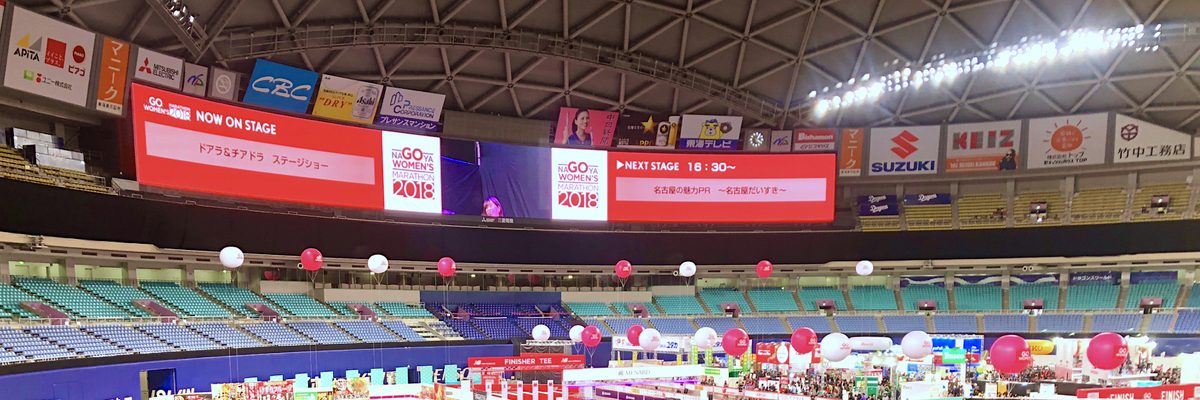 名古屋巨蛋內的馬拉松 EXPO:圓頂下方掛著「NAGOYA WOMEN'S MARATHON 2018」的大型電子看板,看台下的場地搭滿攤位、飄著紅白氣球,左下有一塊「FINISHER TEE」招牌