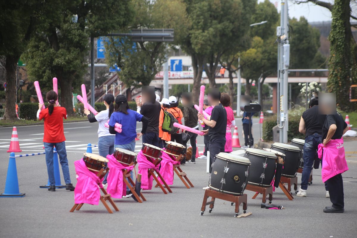 馬拉松當天封路的大馬路邊,一排太鼓架在木架上、披著粉紅色布,應援團手拿粉紅色加油棒等跑者經過,路面放著三角錐