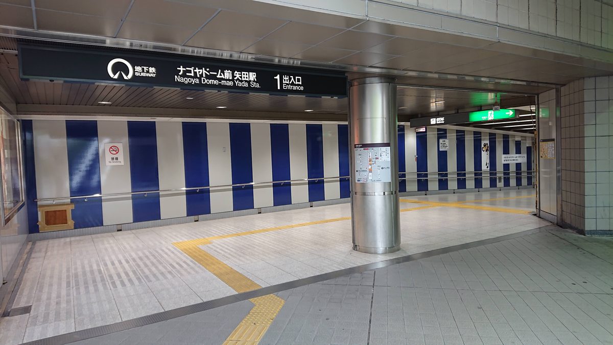 地下鐵ナゴヤドーム前矢田站的地下通道,頭頂黑底白字招牌寫「ナゴヤドーム前 矢田駅 1 出入口」,牆面是藍白相間的直條紋,地上有黃色導盲磚,無人