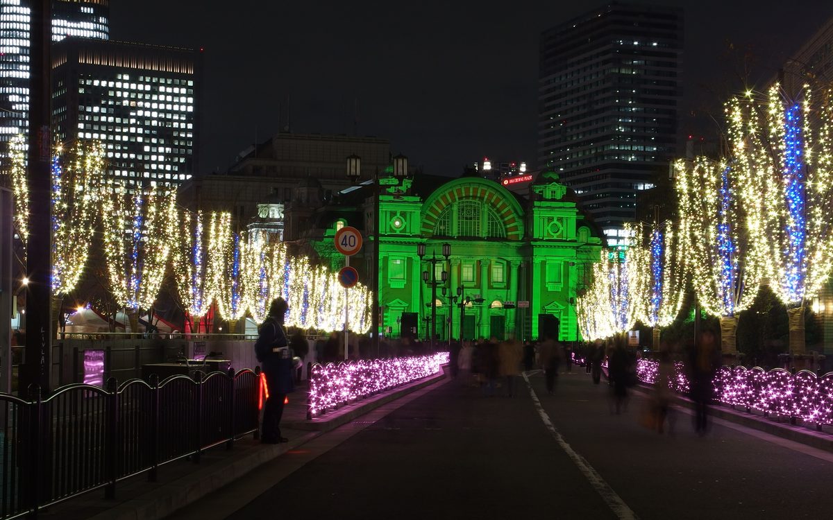 2014 年 12 月夜晚,從東側的路上正對大阪市中央公會堂:公會堂的拱窗正面被打成綠色,路兩旁的行道樹掛著金黃與藍色燈串,路緣是一排粉紅色燈飾,左側樹下看得到幾頂白色帳篷,路上的行人因長時間曝光而模糊