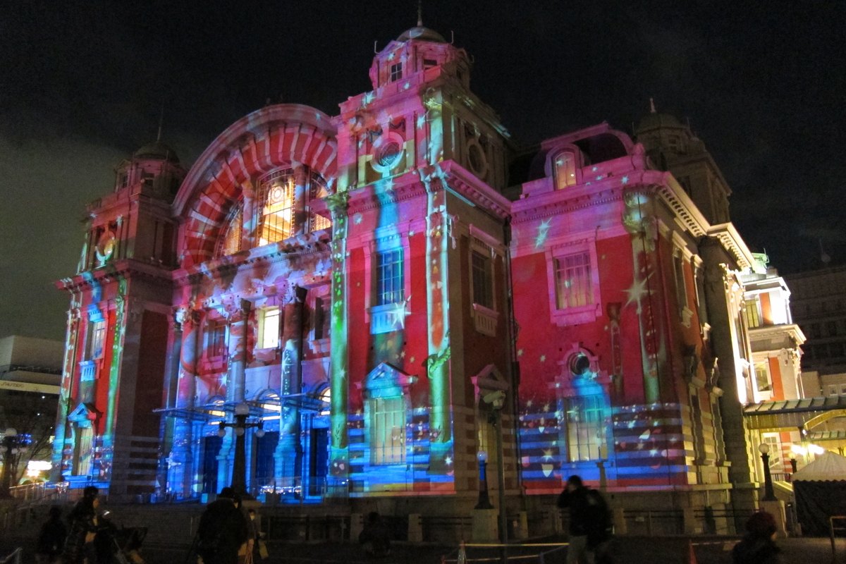 2010 年 12 月夜晚的大阪市中央公會堂:紅磚建築的大拱窗正面被投影成藍、粉紅與綠色的圖案,有星星與緞帶花樣,右下角有一頂白色帳篷,前方是幾個看投影的人的黑色背影