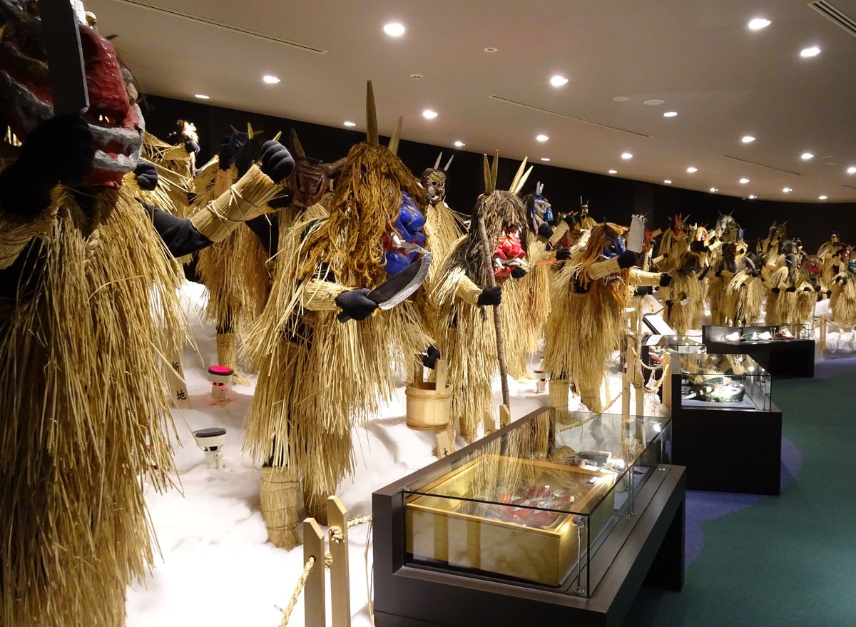 なまはげ館室內展示廳:地面舖著白色人造雪,數十尊等身大的生剝鬼裝束沿著左側牆面一路排到展廳深處,每一尊的面具造型都不同——有紅面尖角、藍面、褐面獠牙、白面等,蓑衣的稻草長短與顏色也各異;人造雪地上插著幾面寫有聚落名的木牌,右側是幾座玻璃展示櫃與深綠色地毯