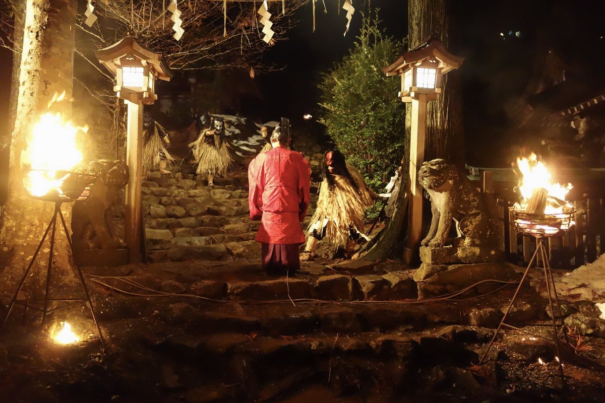 夜間的真山神社參道石階:畫面正中是一段粗石砌的階梯,兩側各立著一座木造的方形石燈籠與一尊石獅子,階梯上方橫掛著垂了白色紙垂的注連繩;左右兩邊各架著一座鐵製三腳火盆,盆裡的柴火燒得正旺,一名穿紅色狩衣、戴烏帽子的神職人員背對鏡頭站在階梯前,階梯上有兩名披稻草蓑衣、戴鬼面的生剝鬼正走下來,畫面右緣的地面留著一小片殘雪