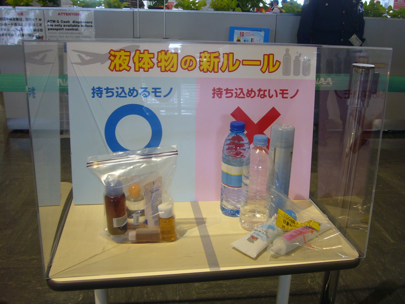 A perspex display case at Narita Airport. The backing board is split down the middle: the left half is pale blue, headed "things you can bring on board" with a large blue circle; the right half is pink, headed "things you cannot" with a red cross. On the left sits a clear zip-lock bag holding several small bottles; on the right stand a capped mineral water bottle, a clear plastic bottle and a tall aerosol can, with two toothpaste tubes lying in front carrying yellow tags.