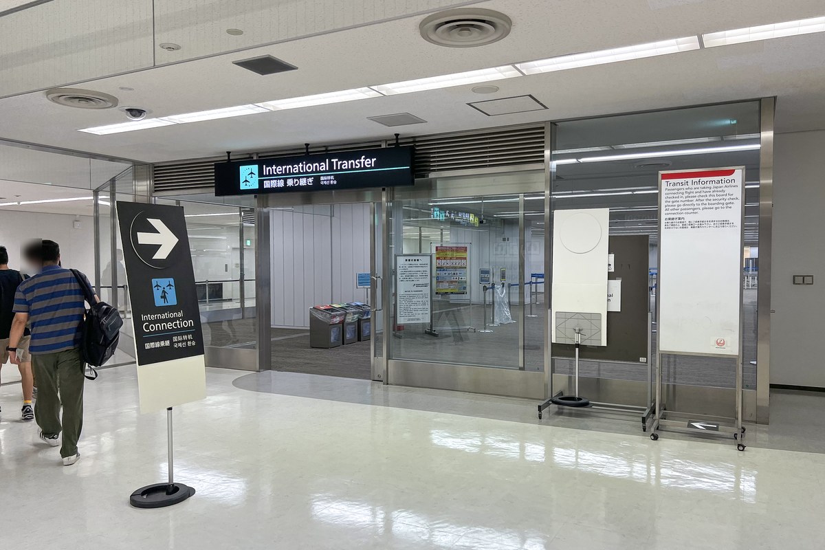 成田機場第 2 航廈下機後的國際線轉機入口：玻璃門上方燈箱寫「International Transfer 国際線 乗り継ぎ」，左邊立牌是箭頭加「International Connection 国際線乗継」，右邊日本航空的 Transit Information 告示板，旅客從左側背對鏡頭走過
