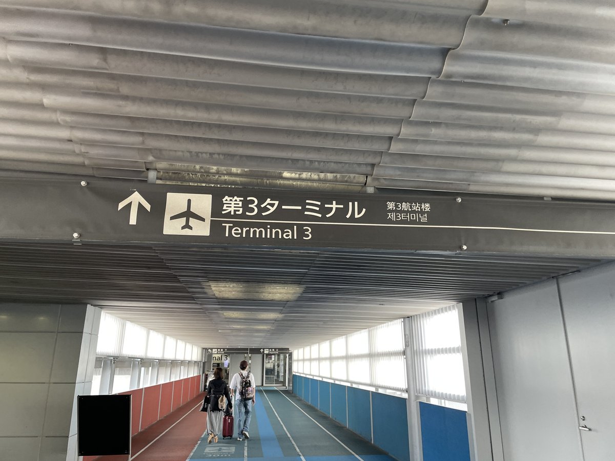 往第 3 航廈的連絡通道:上方指標寫「↑ 第3ターミナル Terminal 3」與中文「第3航站楼」,通道地板中間是藍色、左側是紅褐色,牆面左紅右藍,兩名拖行李的旅客背影往前走