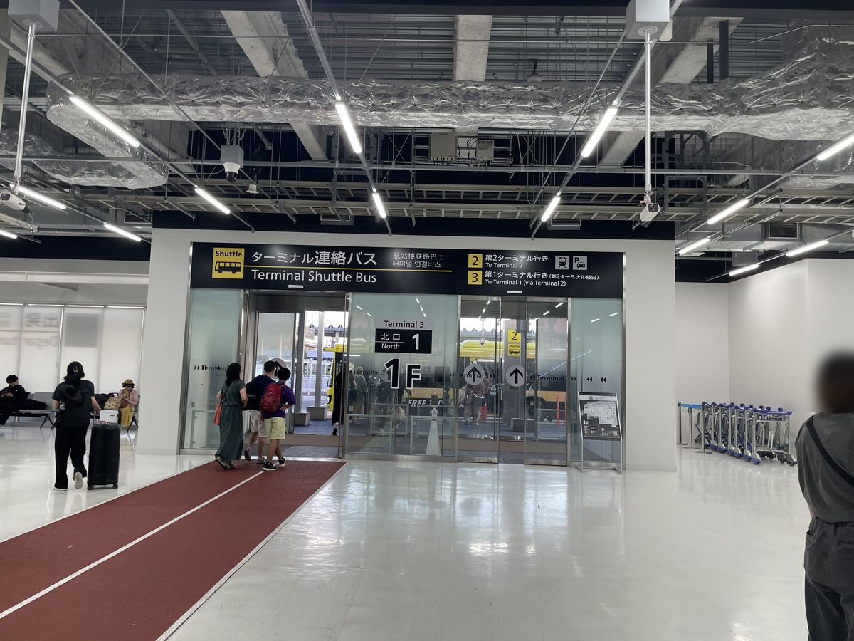 第 3 航廈 1 樓的玻璃出口,上方黑色看板寫「ターミナル連絡バス Terminal Shuttle Bus」,右半寫「2 第2ターミナル行き」「3 第1ターミナル行き(第2ターミナル経由)」,門上標「北口 1 North」「1F」,門外看得到黃色巴士;地上一條紅色通道通往出口,幾位旅客背影走向門口