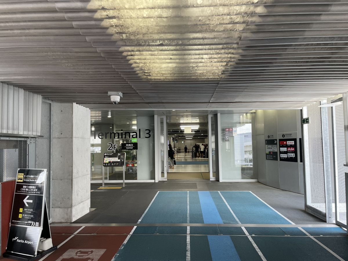 連絡通道盡頭的第 3 航廈 2 樓入口,玻璃門上寫「Terminal 3」「2F」與「中央口 Central」,地板是藍綠色通道;左前方立牌寫連絡巴士乘車處在前方 1 樓,2 號往第 2 航廈、3 號往第 1・第 2 航廈;右側牆上是列出各航空公司標誌的看板