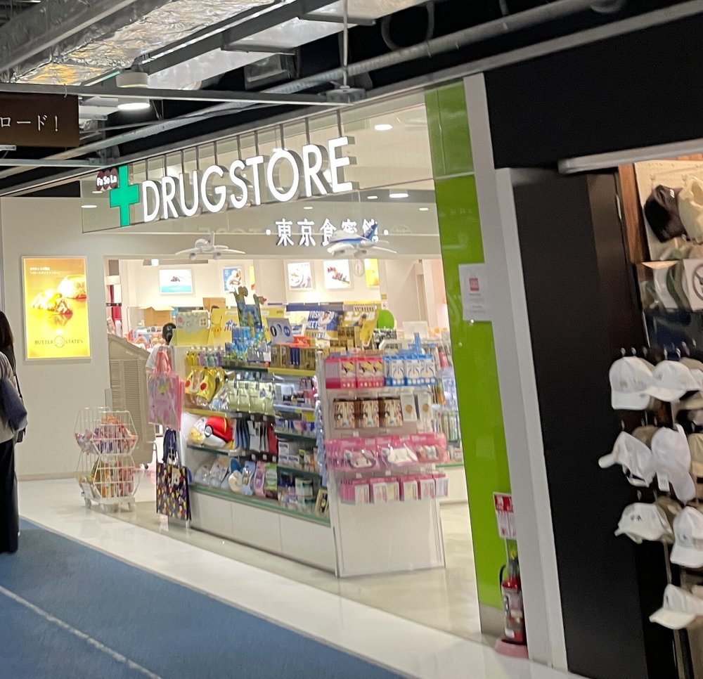 第 3 航廈 2 樓出發大廳旁的店面,白色招牌寫「DRUGSTORE」與「東京食賓館」,店內貨架擺滿零食禮盒、寶可夢周邊與粉紅色包裝的伴手禮,右側是掛滿白色帽子的商店,地上是藍色通道
