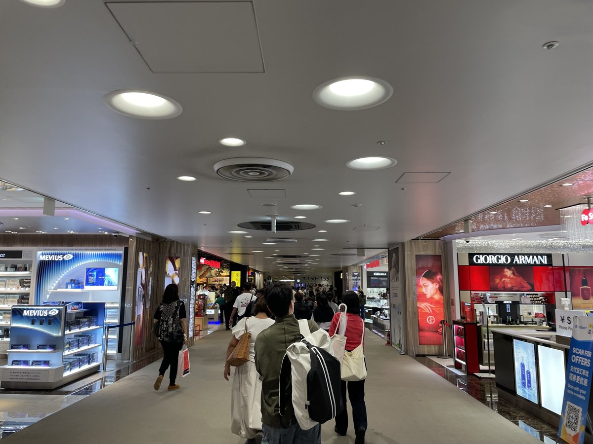 第 3 航廈出境審查後的免稅店走道,左邊是 MEVIUS 香菸的藍色展示櫃,右邊是 GIORGIO ARMANI 化妝品櫃與 Fa-So-La 標誌,走道盡頭的指標寫登機門「150–155」,旅客背影往前走