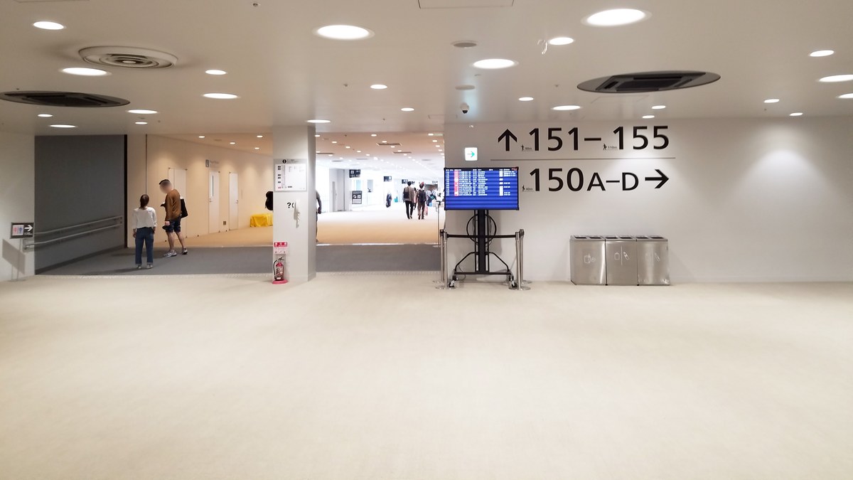 成田機場第 3 航廈出境後的管制區通道：白色地板與天花板圓形燈，右側牆上指標寫「151–155」往前、「150A–D」往右，中間立著一台航班資訊螢幕，遠處有幾位旅客