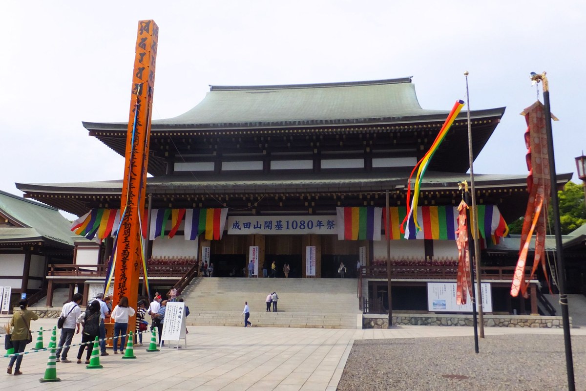 成田山新勝寺大本堂：綠色銅瓦的大屋頂與朱紅梁柱，正面石階前掛著五色幕與「成田山開基1080年祭」橫幅，左側立著寫滿墨字的橘色高柱，廣場上有參拜者