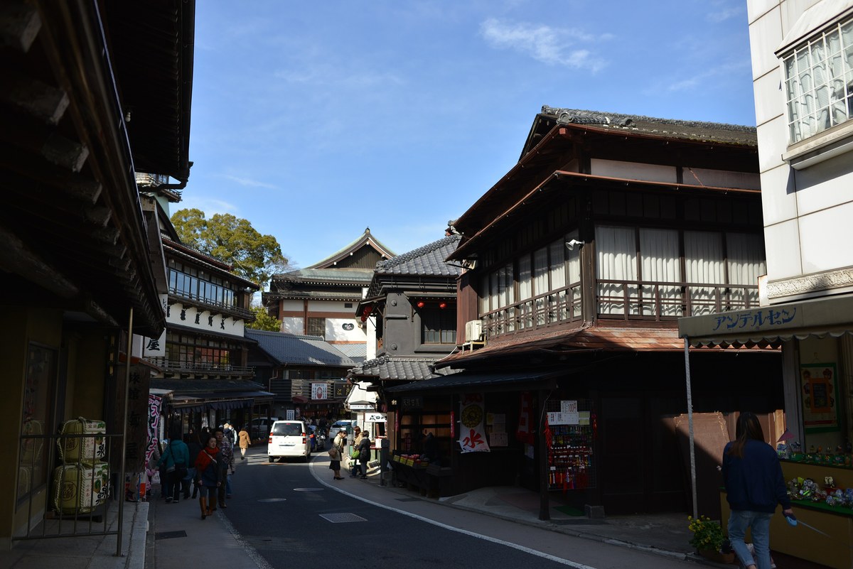 成田山表參道的彎曲坡道：兩側是木造二層老店鋪，盡頭露出寺院的屋頂，路上有零星行人與一台白色廂型車