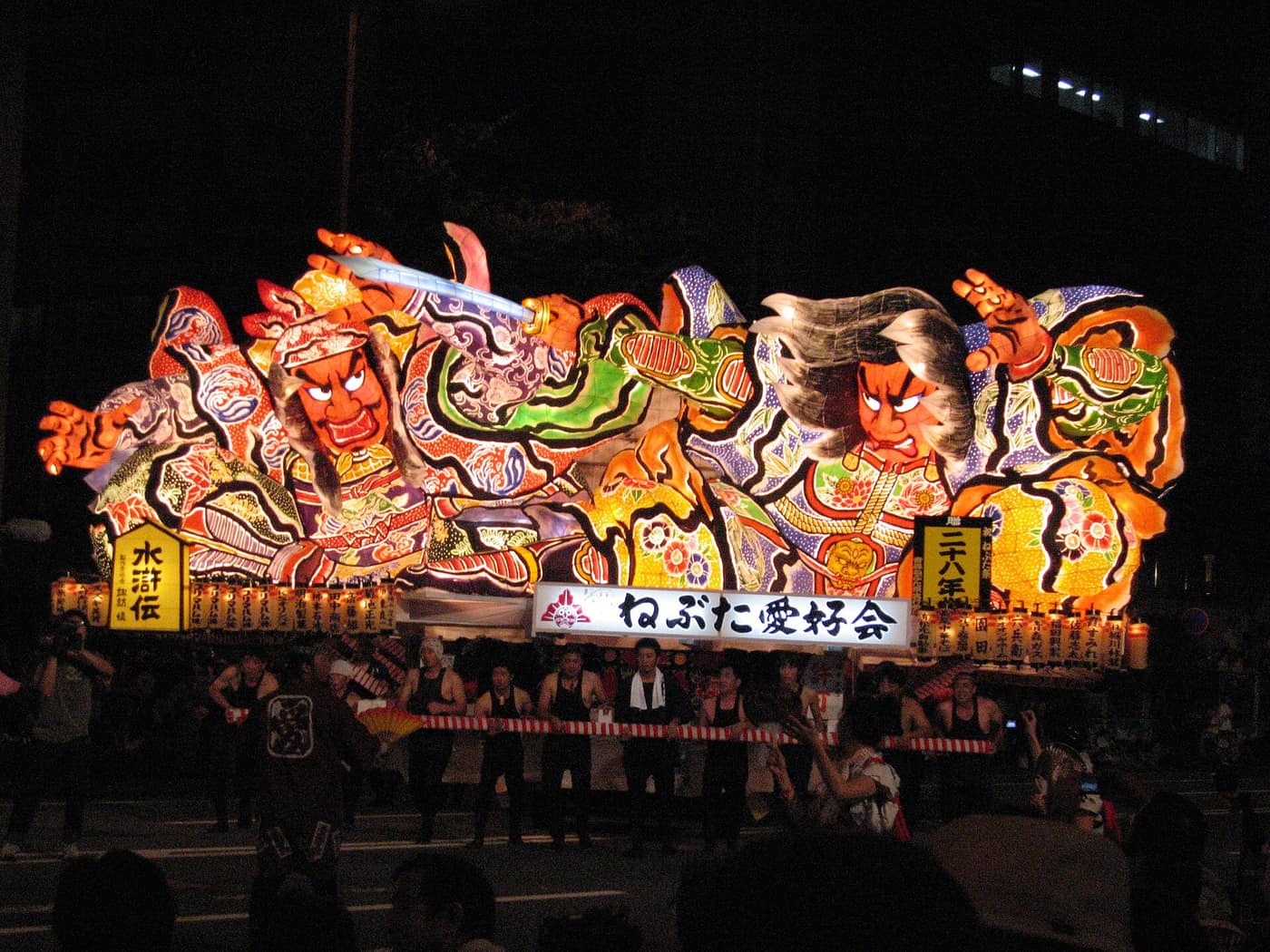 路邊視角看青森睡魔祭夜間運行，大型睡魔下方一排曳き手扶著紅白橫桿前進