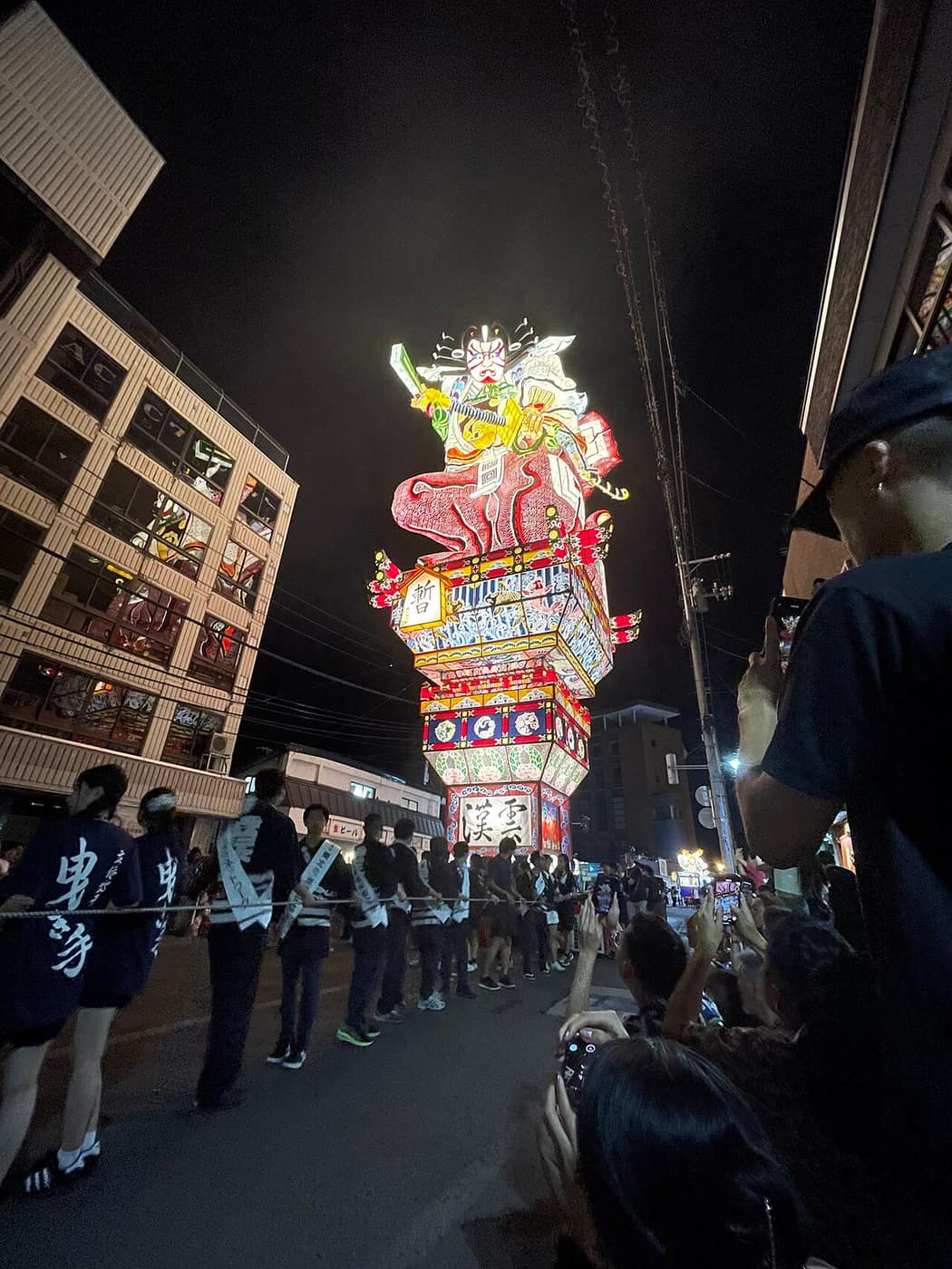 五所川原立佞武多祭典中直立的巨型燈籠山車在夜間街道運行，兩側是曳き手與觀眾