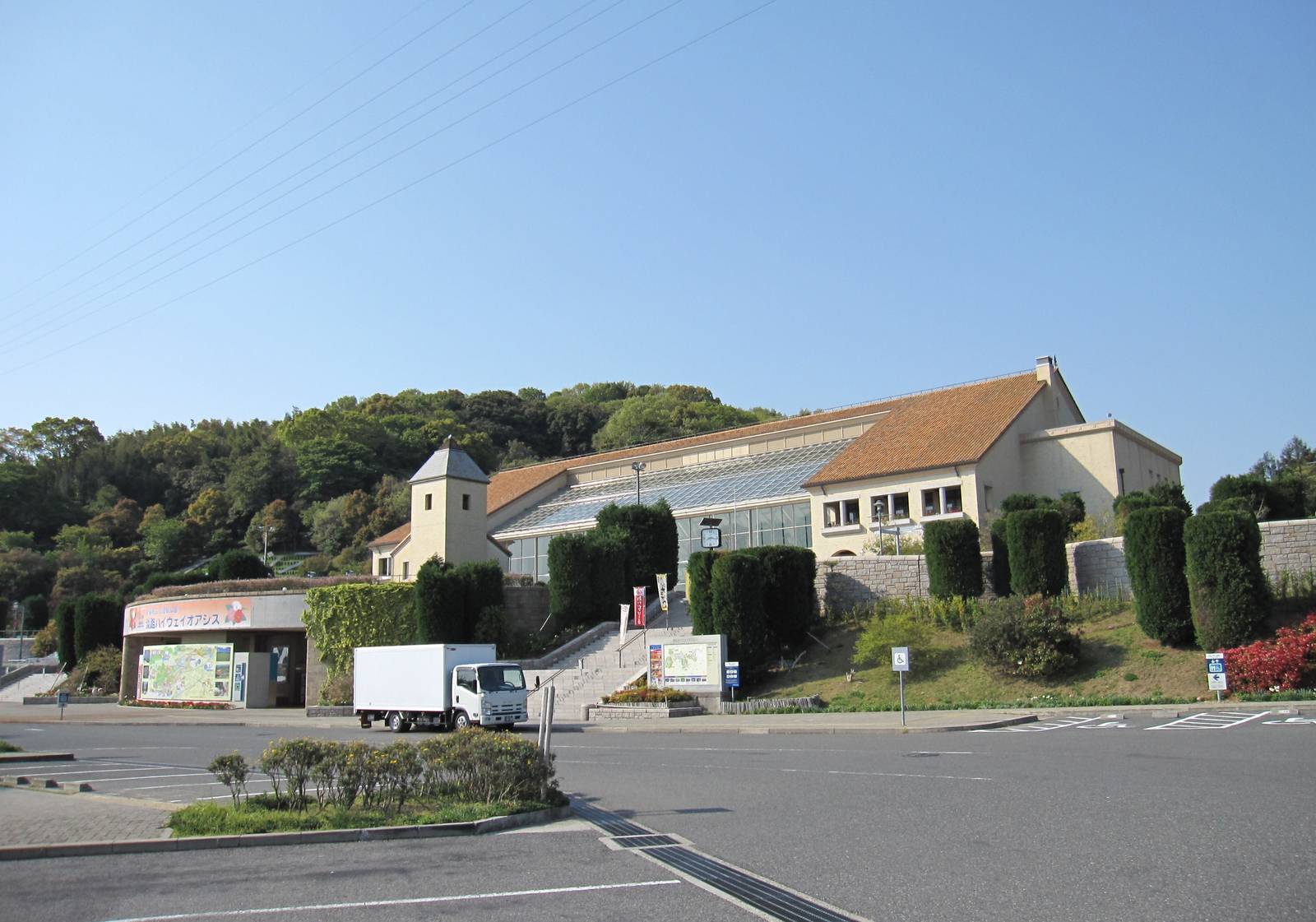 淡路ハイウェイオアシス的建築:橘色屋頂與玻璃屋頂的米色建物蓋在山坡上,前方停車場旁有階梯往上,左側入口掛著「淡路ハイウェイオアシス」招牌