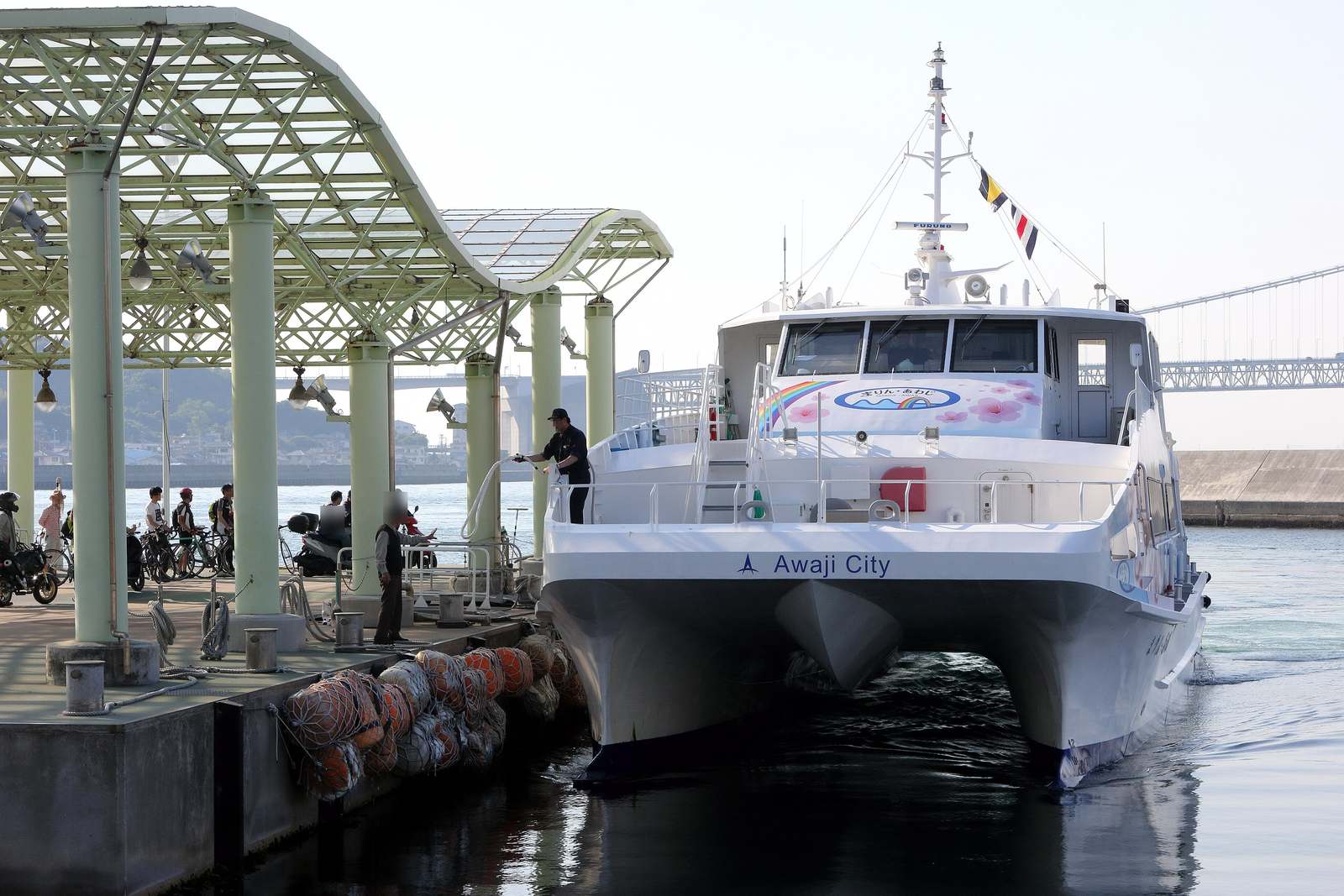 淡路ジェノバライン的白色雙體高速船「まりん・あわじ」靠上岩屋港的棧橋,船員在甲板上拋纜繩,棧橋淺綠色柱子後面排著等著上船的自行車和機車騎士