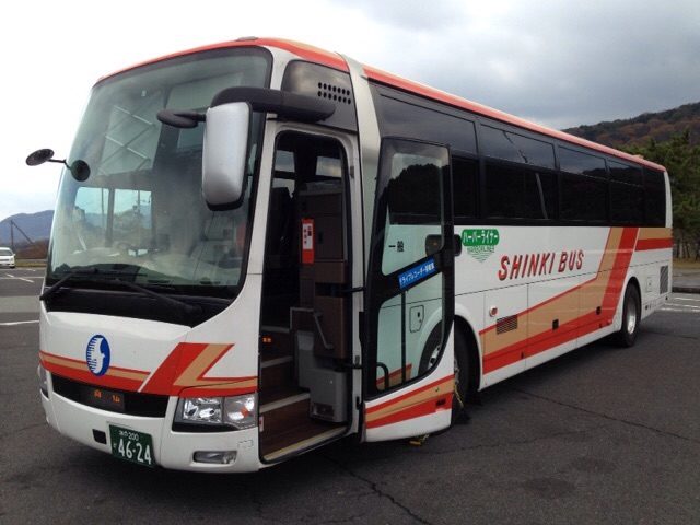 一輛白色車身、側面橘紅色斜條紋、寫著 SHINKI BUS 的神姬巴士高速巴士,前門打開停在停車場上