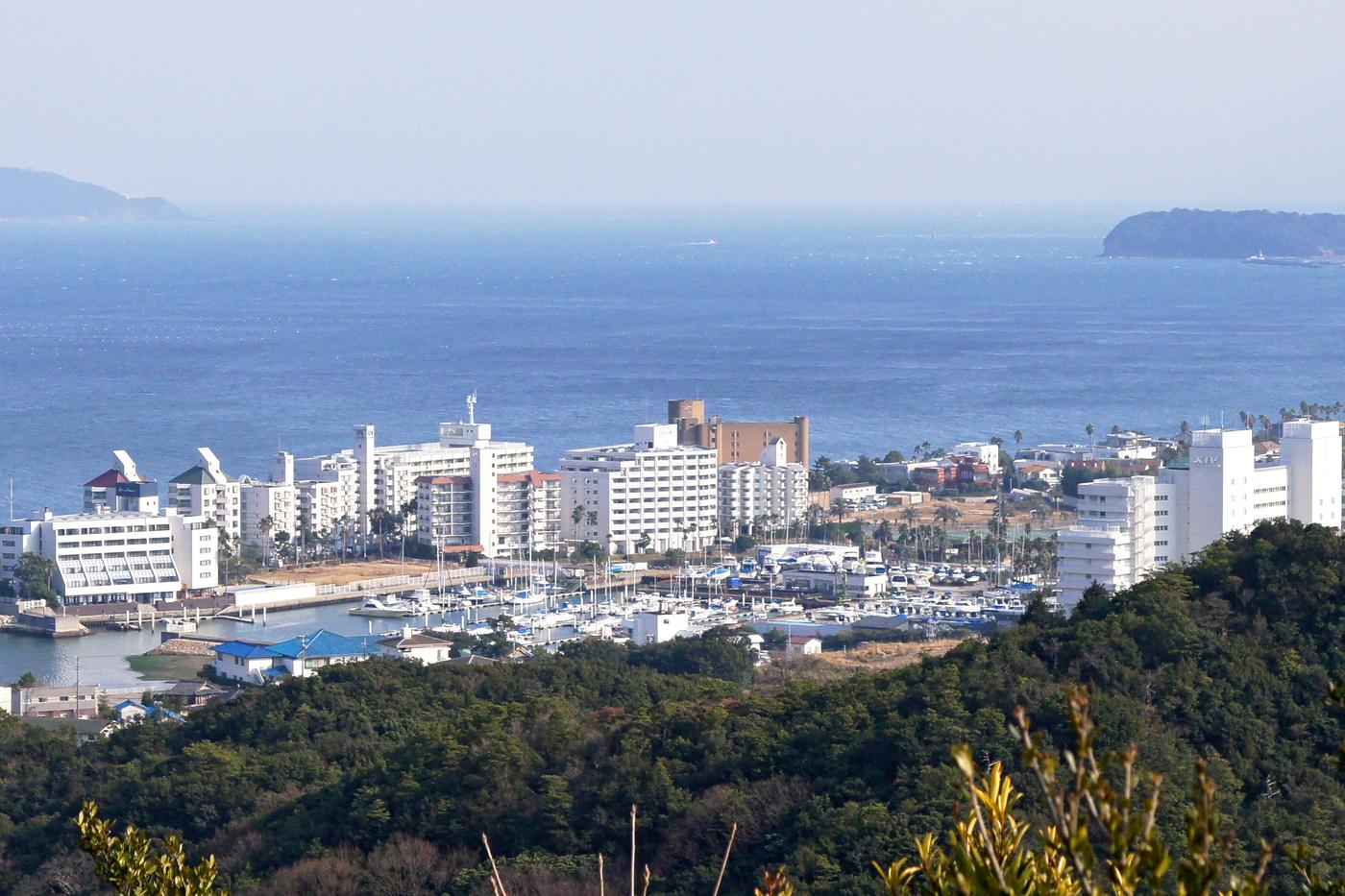 從山上俯瞰洲本溫泉:前景是綠色山林,中間沿著海岸排著一整列白色飯店大樓與遊艇碼頭,後方是一片大阪灣海面
