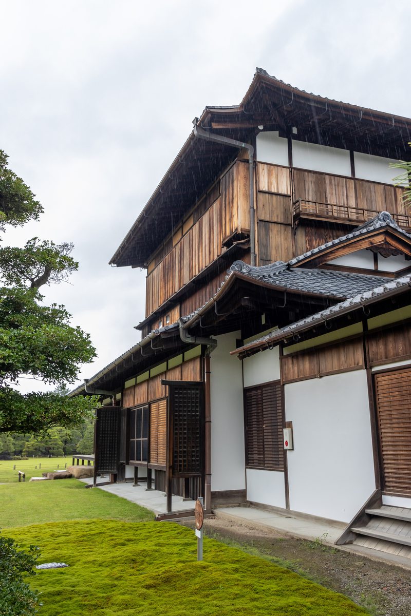 二條城本丸御殿的一棟多層木造建築側面:上層是深褐色木板牆與木格窗,下層是白牆與木板雨戶,屋簷下有木造階梯,建築前方是青苔與草坪