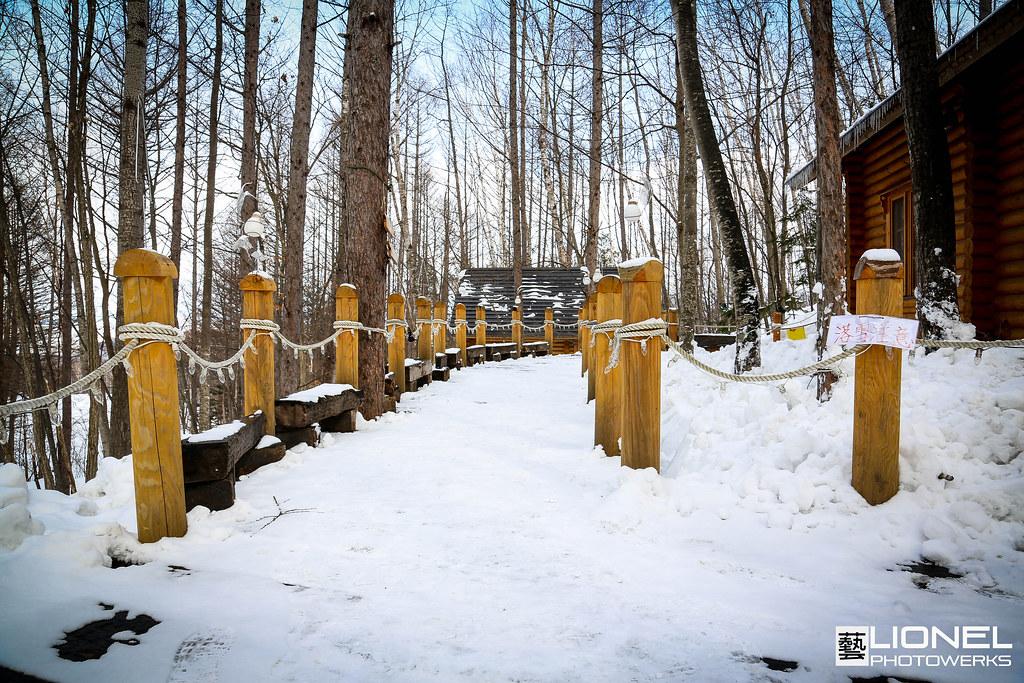 森林裡一條被雪蓋住的木棧道,兩側是一根根纏著粗麻繩的木樁與木頭長椅,右邊露出原木小屋的一角,繩子上掛著一張手寫的紙牌
