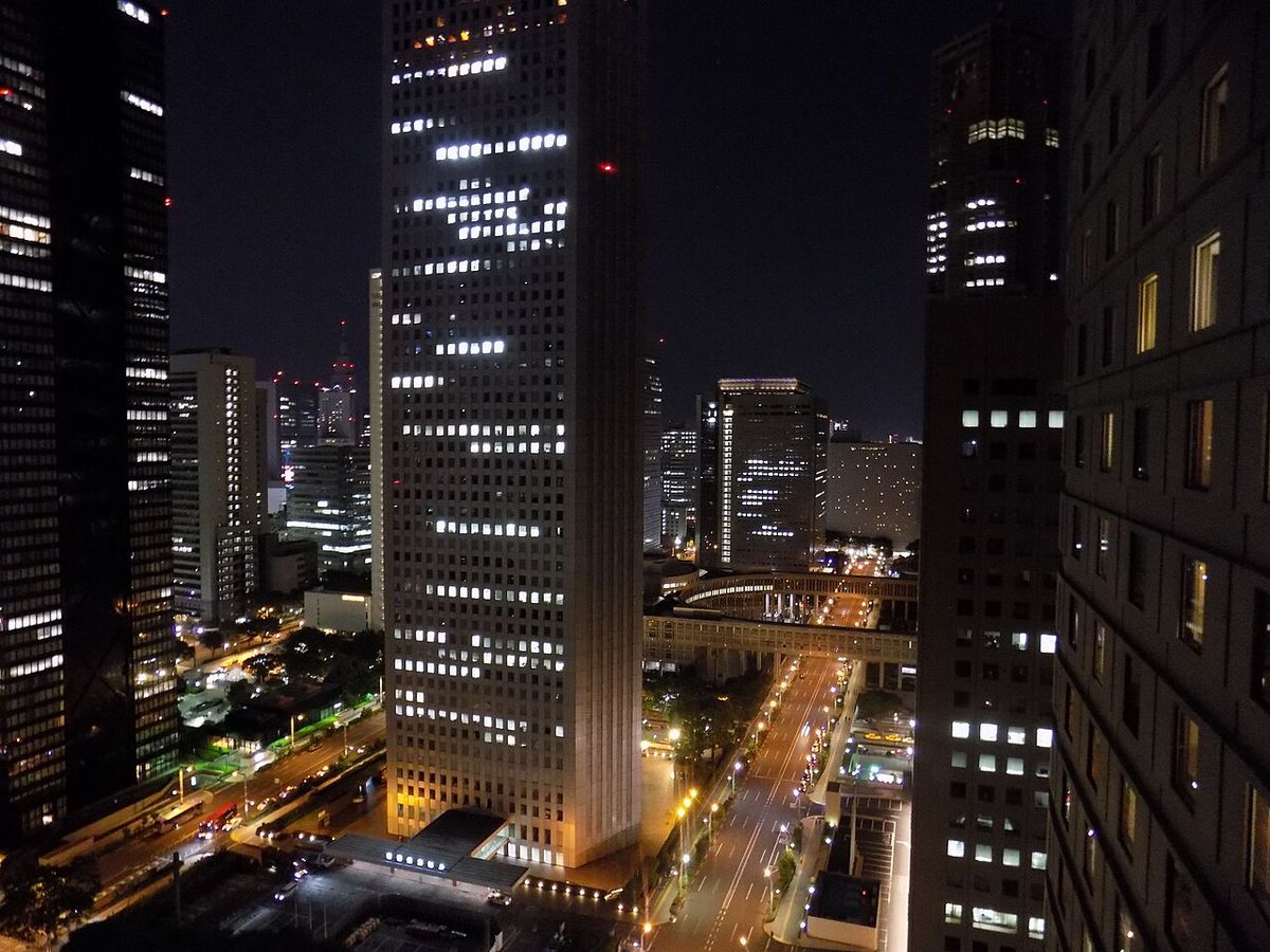 Night view over the Nishi-Shinjuku high-rise district from above: a pale skyscraper with a grid of lit windows in the centre, darker towers on both sides, and a road strung with car and street lights running past a curved pedestrian bridge