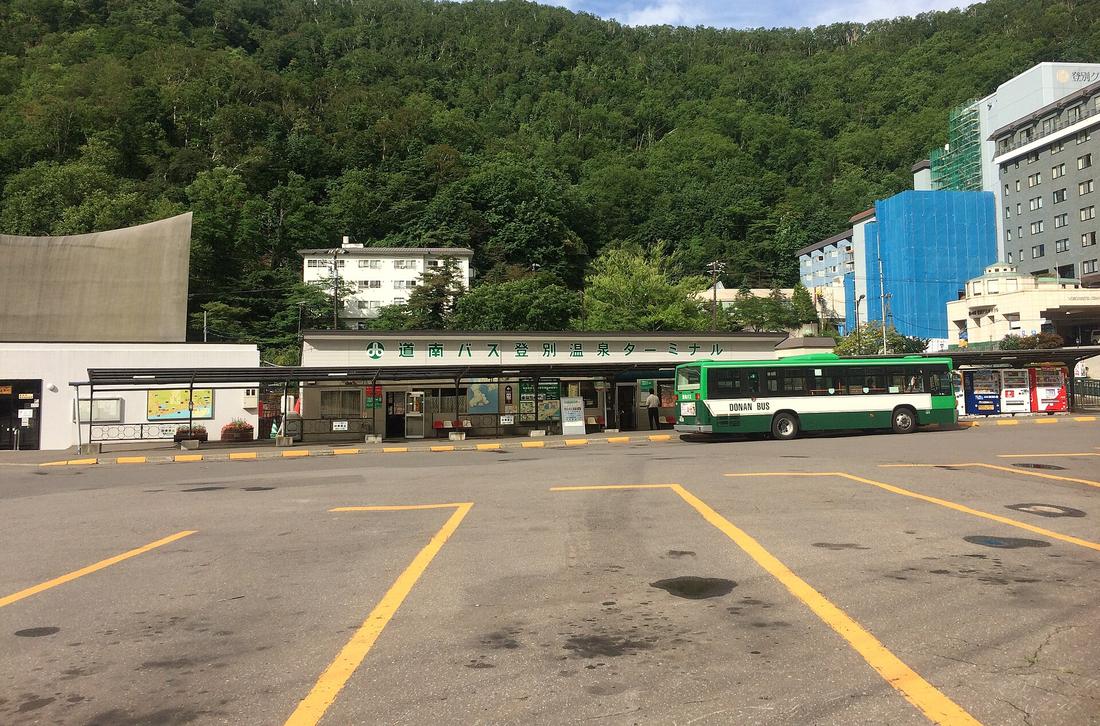 道南巴士登別溫泉總站：低矮的站房招牌寫「道南バス登別温泉ターミナル」，一台綠白色道南巴士停在站前，背後是綠色山坡與溫泉飯店
