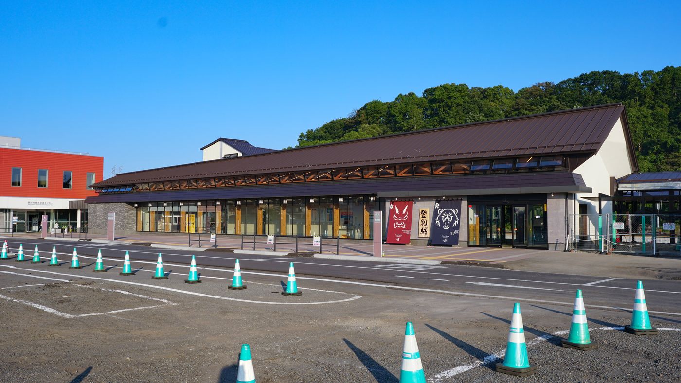 JR 登別站新站舍：深褐色長斜屋頂、整排落地玻璃，入口旁掛著紅色鬼臉與深藍熊臉兩面暖簾和「登別駅」木牌；左邊是紅色外牆的登別市觀光交流中心
