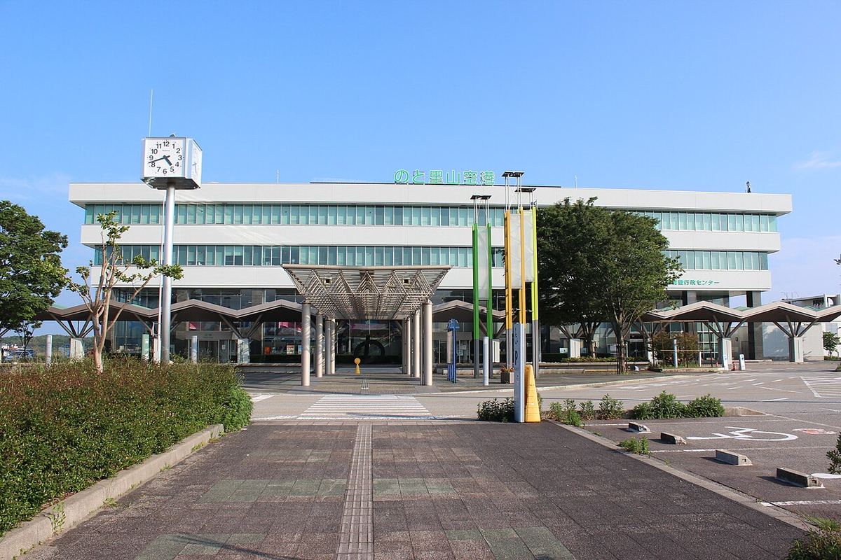 The front of Noto airport: a white three-storey terminal with green lettering reading Noto Satoyama Airport, a four-faced clock tower to the left, a canopy and three flagpoles at the entrance, and a completely empty set-down lane and car park in front