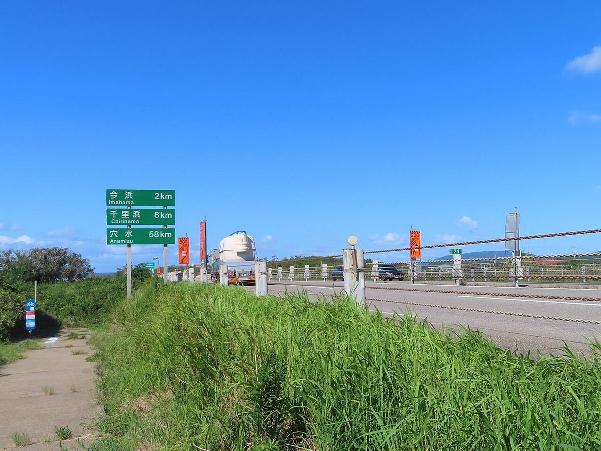 A green distance sign beside the Noto Satoyama Kaido reading Imahama 2km, Chirihama 8km and Anamizu 58km, with a tanker truck and cars running on the open carriageway behind it under a cloudless sky
