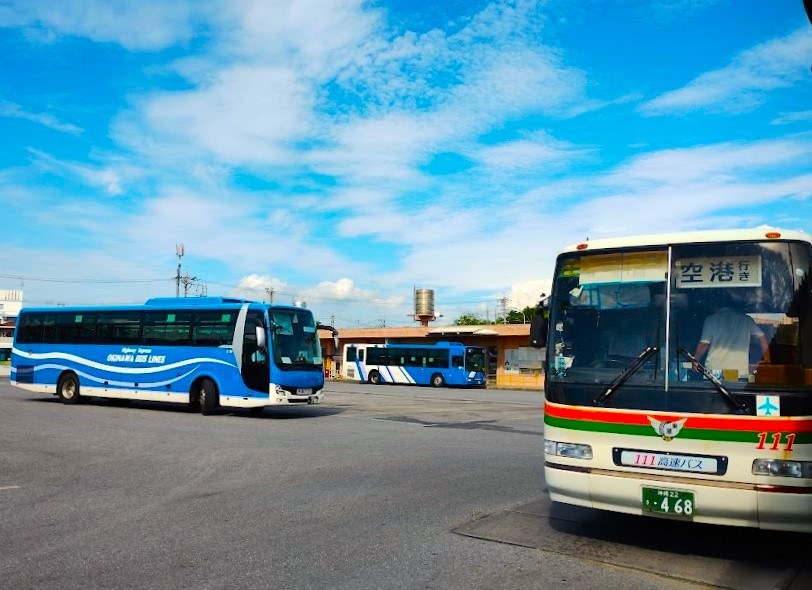 名護巴士總站停車場裡的高速巴士:右邊車頭的行先牌寫「空港行き」,車頭下方標著「111 高速バス」與紅綠色條紋;左邊是藍白色車身寫著 OKINAWA BUS LINES 的巴士