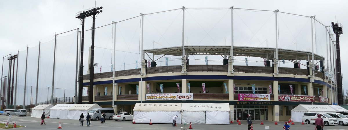 金武町ベースボールスタジアム正面全景:兩層看台外牆掛著「東北楽天ゴールデンイーグルス」的歡迎布條,入口上方是球場名稱,前方廣場搭著金武町教育委員会的白色帳篷,旁邊停了幾輛車