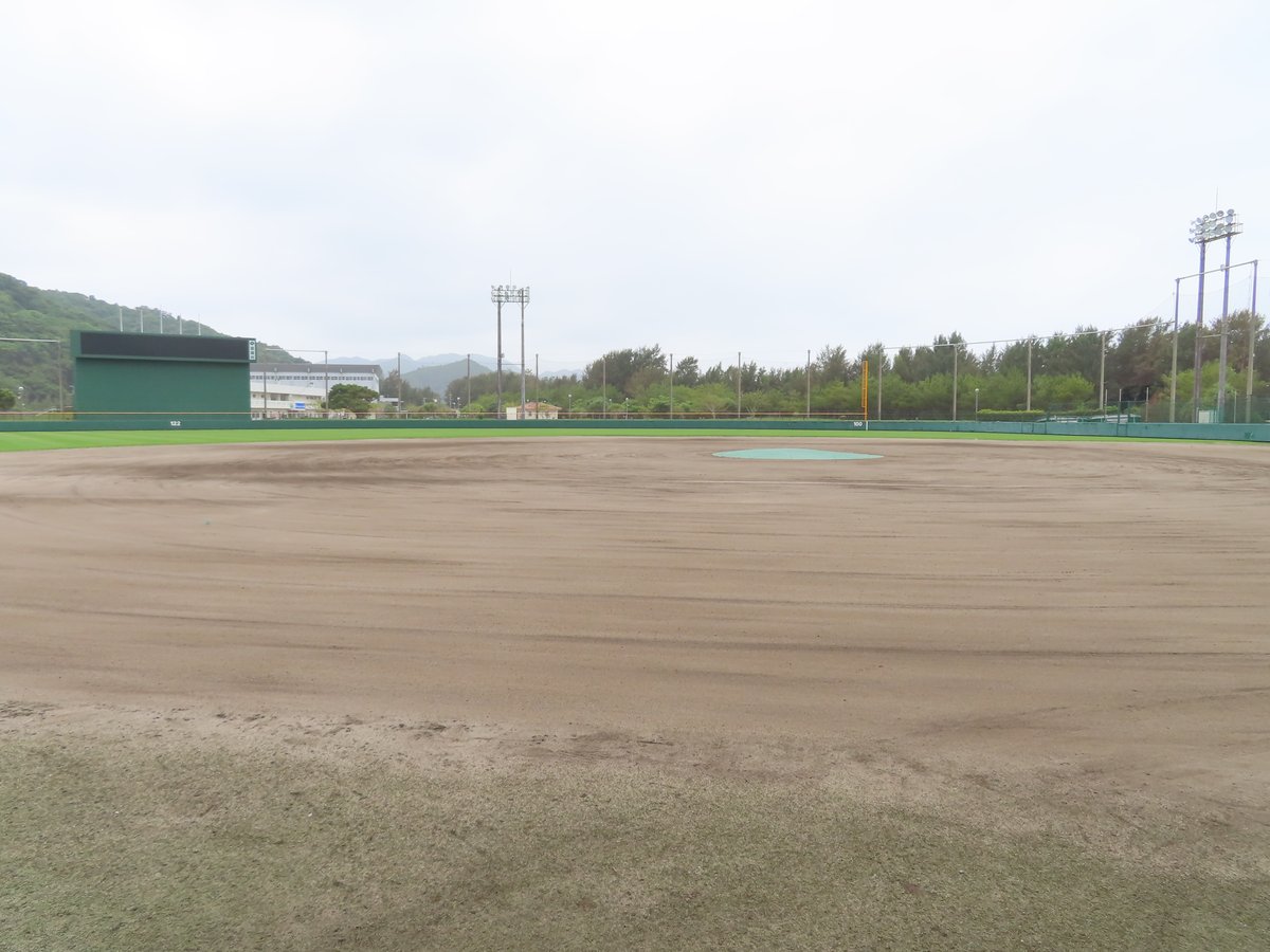 國頭村的くにがみ球場,從本壘後方看出去:整片空的紅土內野與投手丘,外野左側是綠色記分板,後面是山與幾棟建築,右側是照明塔與樹林,天空陰