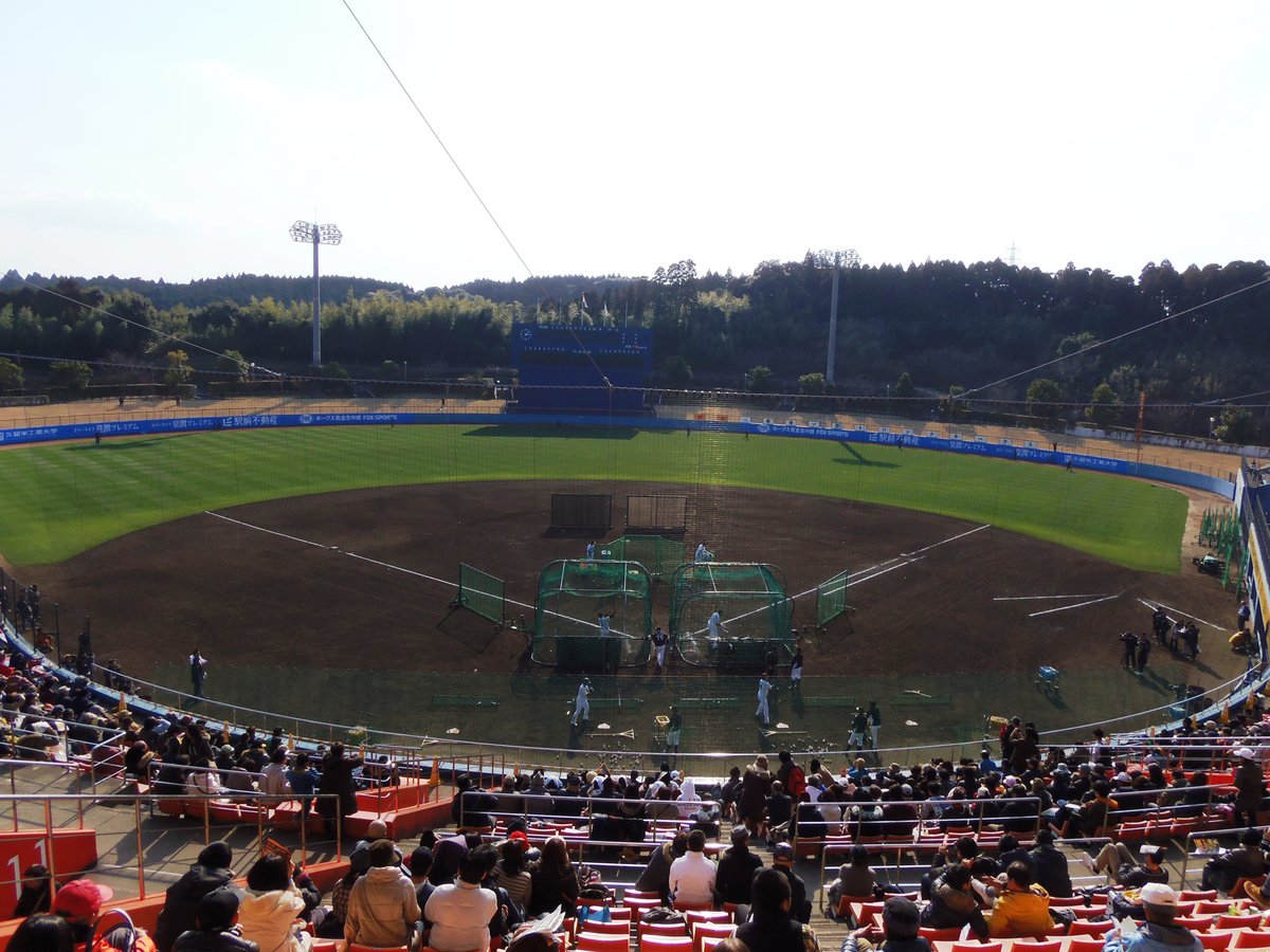 宮崎アイビースタジアム的內野看台坐滿觀眾,大家背對鏡頭看著下方球員在本壘後方的打擊籠練打擊,外野後面是藍色記分板與一整片樹林