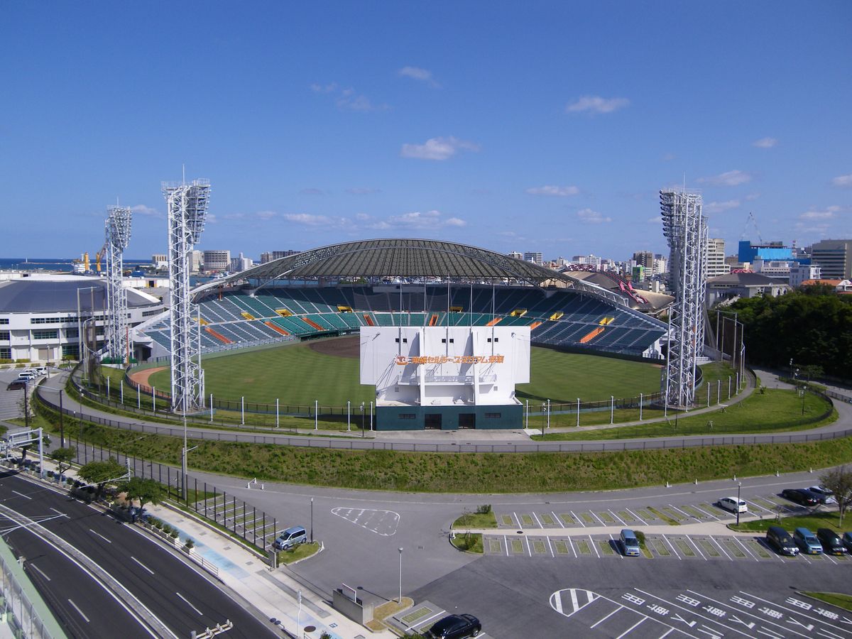 從高處俯瞰沖縄セルラースタジアム那覇:弧形屋頂罩著藍綠色內野看台,草地球場前方是白色記分板背面,左右兩座照明塔,下方是空蕩的停車場與道路,遠方是那霸市街與海