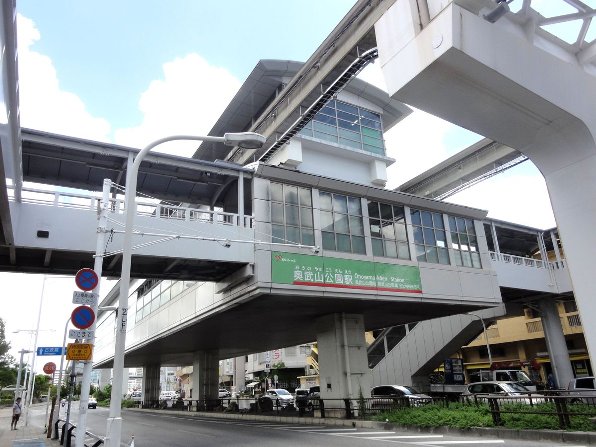 沖繩單軌奥武山公園站的高架站房,綠色站名牌寫「奥武山公園駅 Onoyama-kōen Station」,站房兩側是通往地面的樓梯與天橋,下方是馬路與停著的車