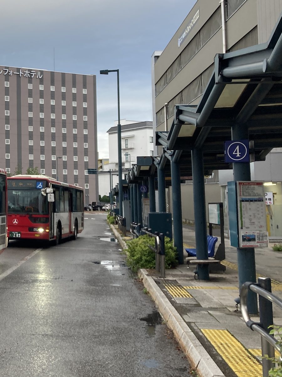 帶廣站巴士總站的候車月台：深藍色鋼柱撐著長雨棚，柱子上掛著藍底白字圓圈「4」號牌和時刻表看板，往後依序是 3 號、2 號；左邊車道上停著紅白色的拓殖巴士