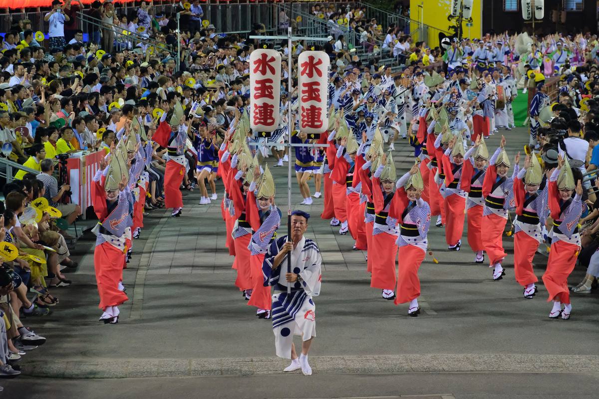Awa Odori women's dance in Tokushima: dancers in yukata and low half-moon straw hats moving on tiptoe in a single file along a floodlit night street between spectator stands