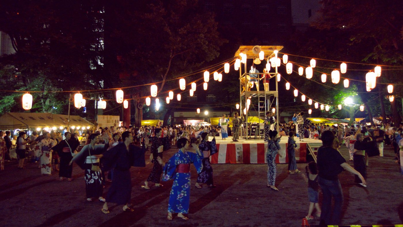 A night-time Bon dance: a wooden yagura tower hung with red and white paper lanterns, taiko drummers on the platform, and a crowd in yukata circling it in slow concentric rings