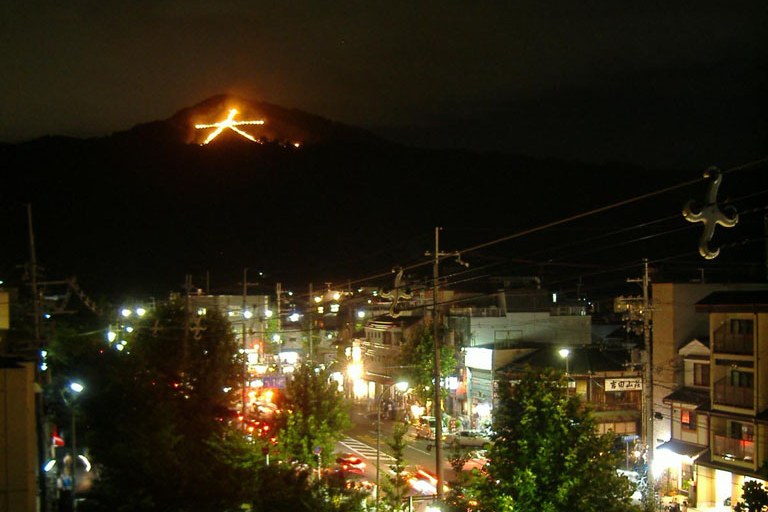 The Daimonji character of the Kyoto Gozan no Okuribi burning on the slope of Mount Nyoigatake at night, seen over the rooftops and scattered lights of the city