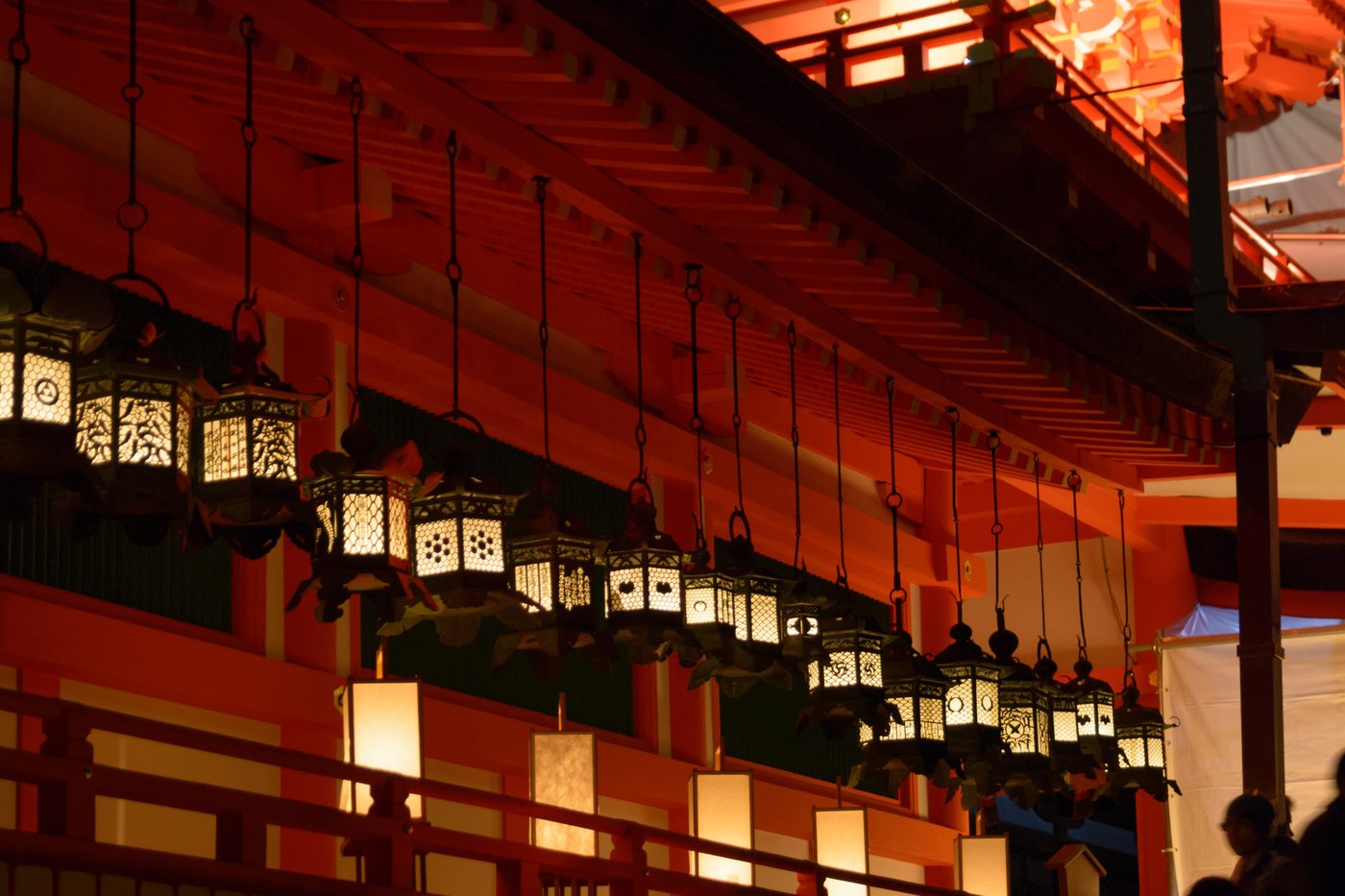 Rows of bronze hanging lanterns lit along the vermilion corridor of Kasuga Taisha at night during the Mantoro, with stone lanterns glowing on the ground beyond