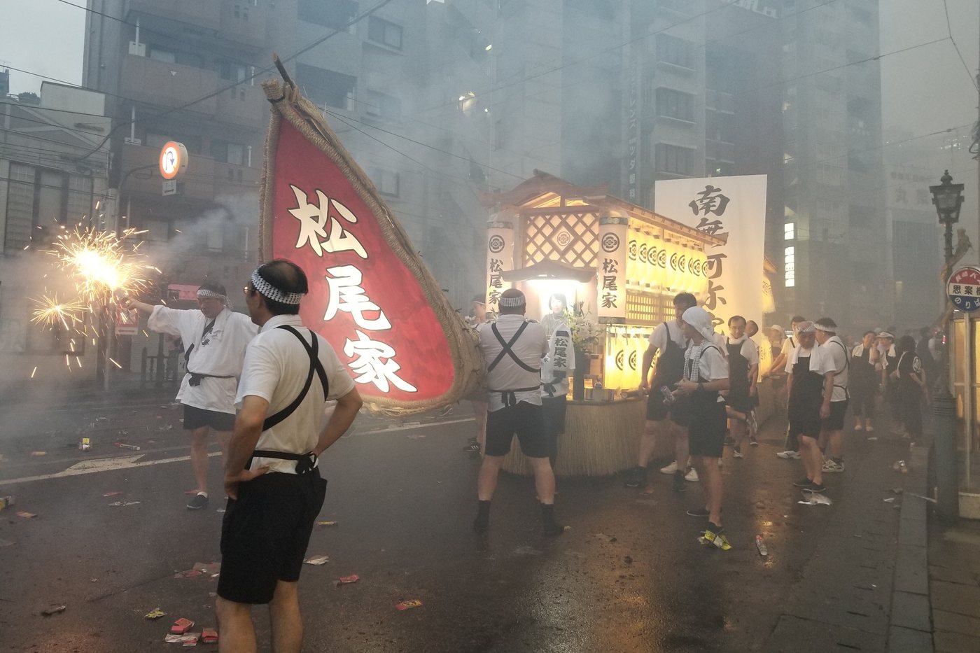 A wooden spirit boat hung with lanterns and paper flowers pushed by family members along a Nagasaki street at night, with spent firecrackers and white smoke across the road ahead