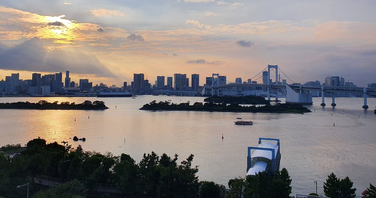 夏天傍晚從 AQUA CiTY 台場往外看的東京灣：夕陽從雲後透出光，海面上有兩座長滿樹的小島，右邊是白色的彩虹橋，對岸是一整排市區高樓，右下角是水上巴士的白色棧橋