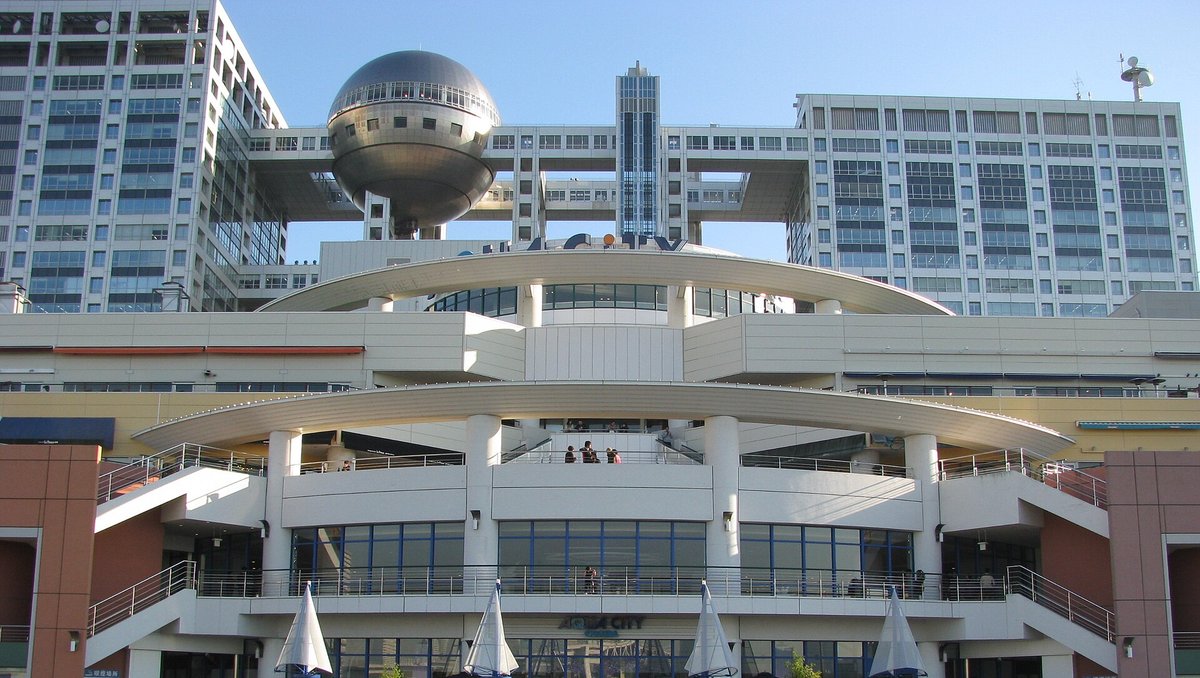 AQUA CiTY 台場面海的那一側：白色的弧形屋簷下有兩層帶欄杆的露台，兩側各有一道戶外樓梯，露台上站著幾個人，建築後方是富士電視台大樓與銀色球體展望室