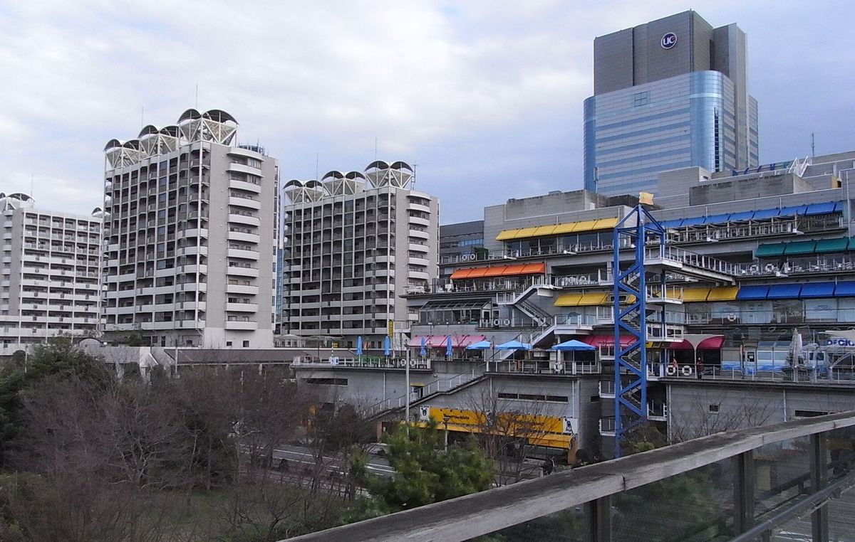 DECKS 東京 Beach 面海側的外觀：灰色建築外側是一層層階梯狀的戶外露台，露台上掛著黃、橘、藍、綠、粉紅色的遮陽棚，中間有一座藍色鋼架樓梯塔，左邊是兩棟高層住宅，前景是木製欄杆