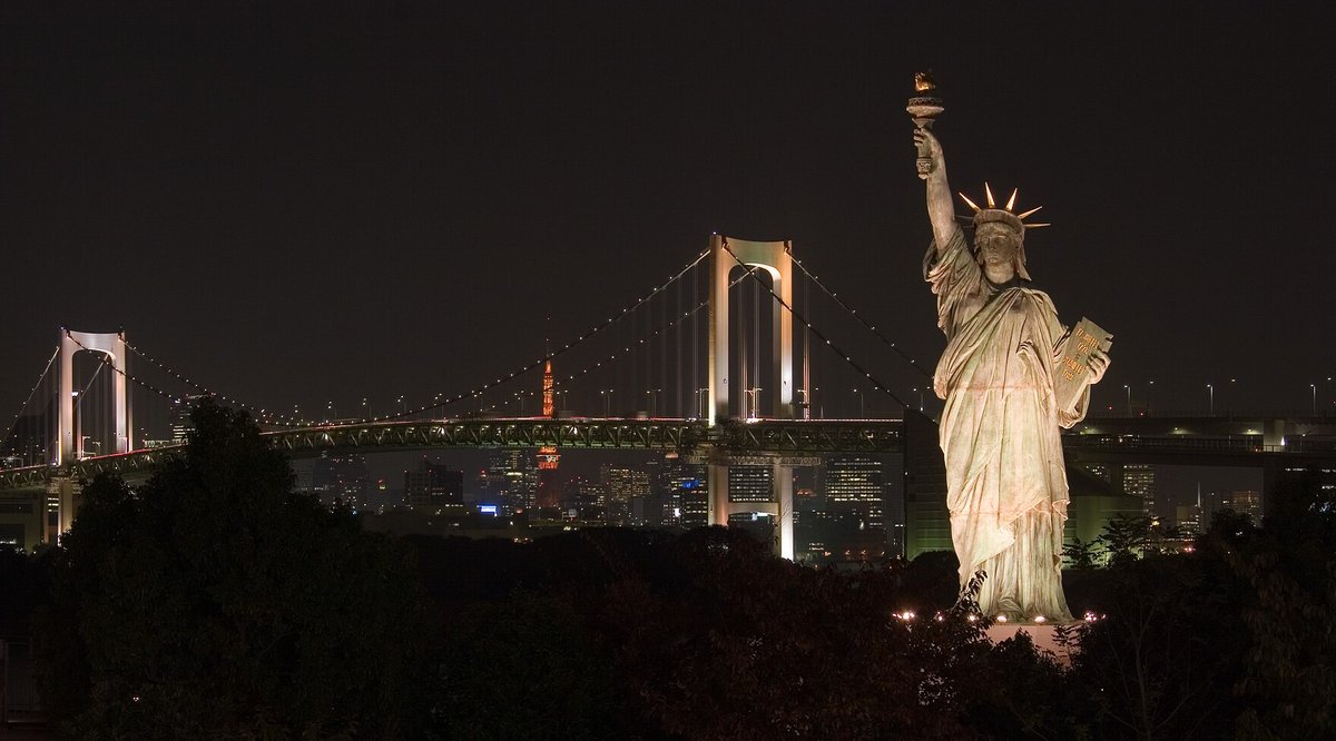 台場的夜景：右邊是打著暖色燈光的自由女神像，左邊是橋塔打白光的彩虹橋，橋的中間露出橘紅色的東京鐵塔，前景是一片黑色的樹影