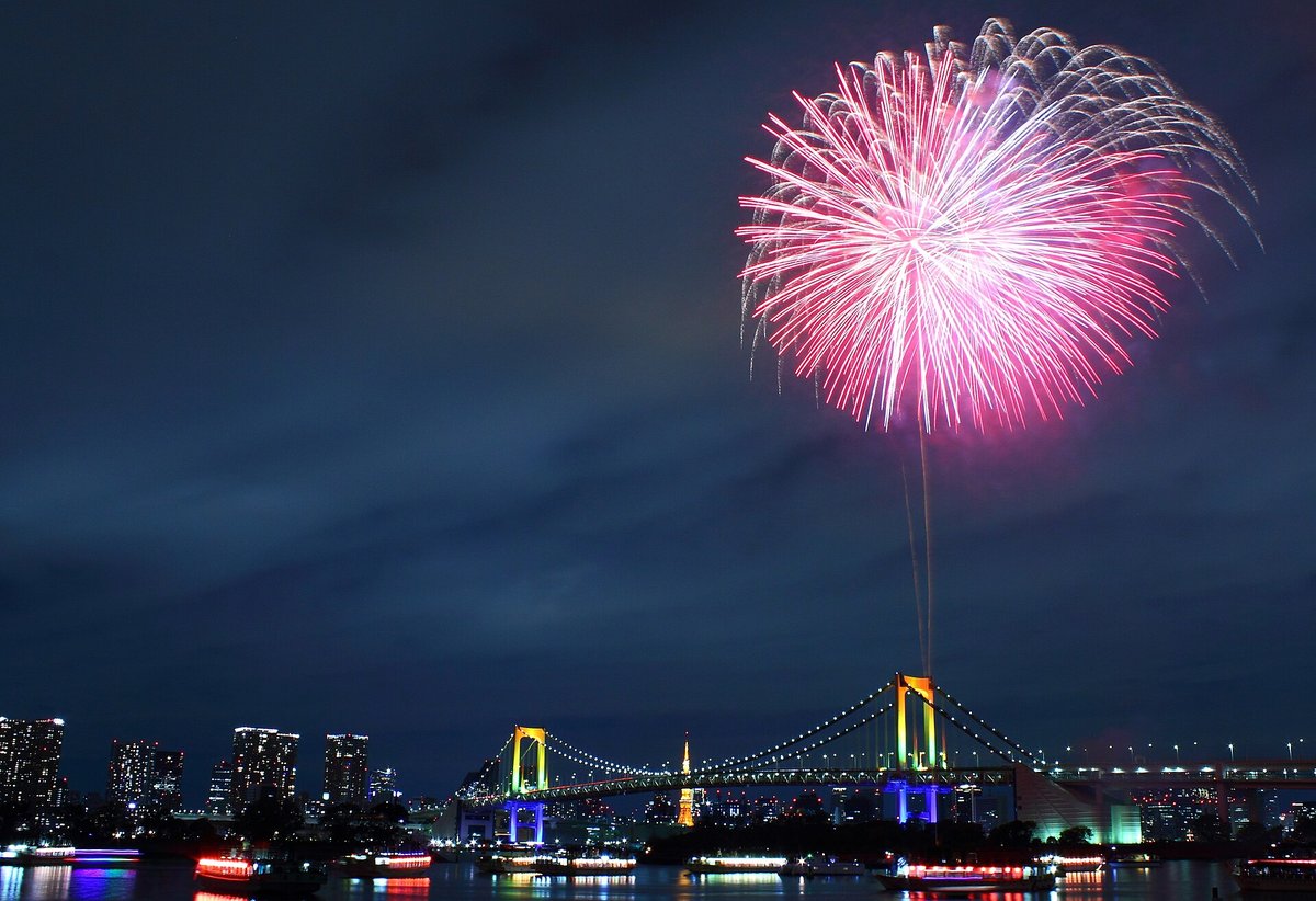 2014 年 12 月的台場彩虹煙火：深藍色夜空裡一朵粉紅色的煙火開在彩虹橋右側橋塔的正上方，橋塔打著黃、綠、藍的燈光，橋後看得到橘色的東京鐵塔，海面上停著一排亮燈的屋形船
