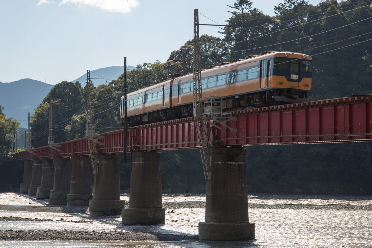 橘色與深藍色的兩節電車開過大井川第一橋梁，紅色鋼梁架在一整排混凝土橋墩上，橋下是寬闊的礫石河床與反光的河水，遠處是山