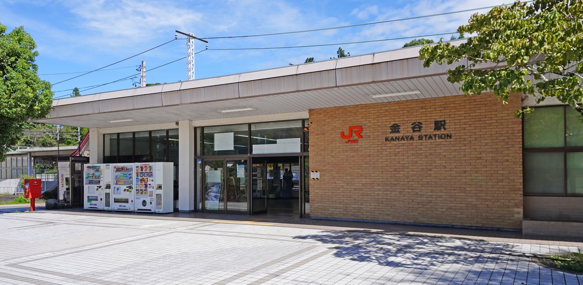 JR 金谷站站舍：平屋頂的一層樓建築，右側磚牆上有橘色 JR 標誌與「金谷駅 KANAYA STATION」字樣，玻璃門左邊有三台自動販賣機與一個紅色郵筒