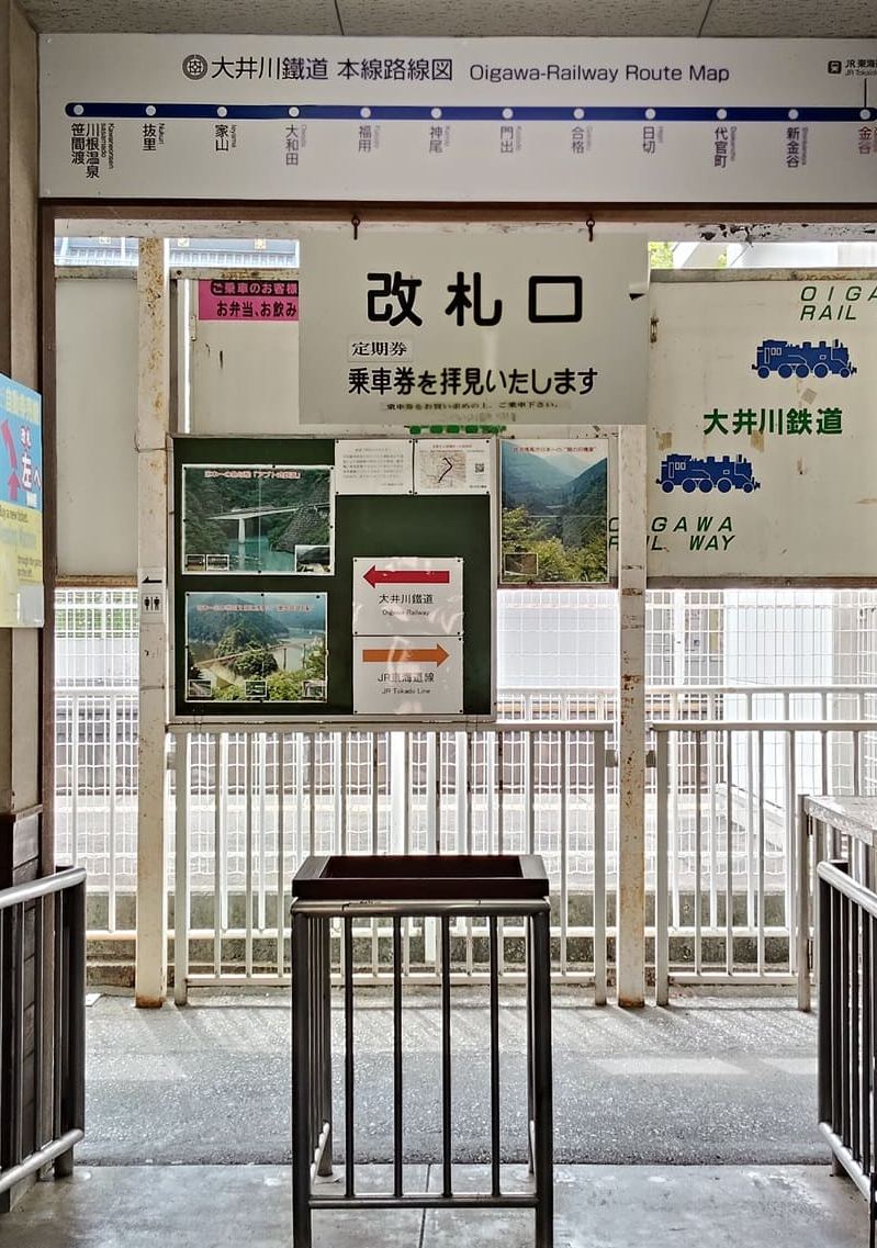 大井川鐵道金谷站的改札口：上方掛著本線路線圖，最左邊一站是川根溫泉笹間渡、最右邊是金谷；下面是「改札口」牌子、不鏽鋼剪票台，綠色看板上有紅色箭頭往左指大井川鐵道、橘色箭頭往右指 JR 東海道線